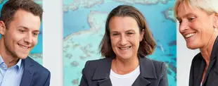 Drei Personen in Business-Kleidung lächeln während eines Gesprächs in einem modernen Büro mit blauen Bildern im Hintergrund.