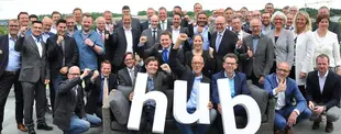 Eine große Gruppe Geschäftsleute posiert freudig auf einer Terrasse, einige halten große Buchstaben, die das Wort „hub“ bilden. Viele zeigen Daumen hoch oder jubeln.