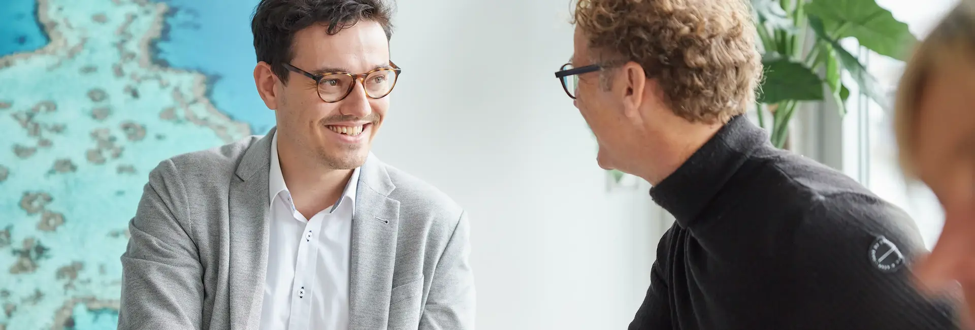 Zwei Männer in einem Gespräch, einer trägt einen grauen Blazer, der andere einen schwarzen Pullover. Sie lachen und wirken engagiert, im Hintergrund eine Pflanze.