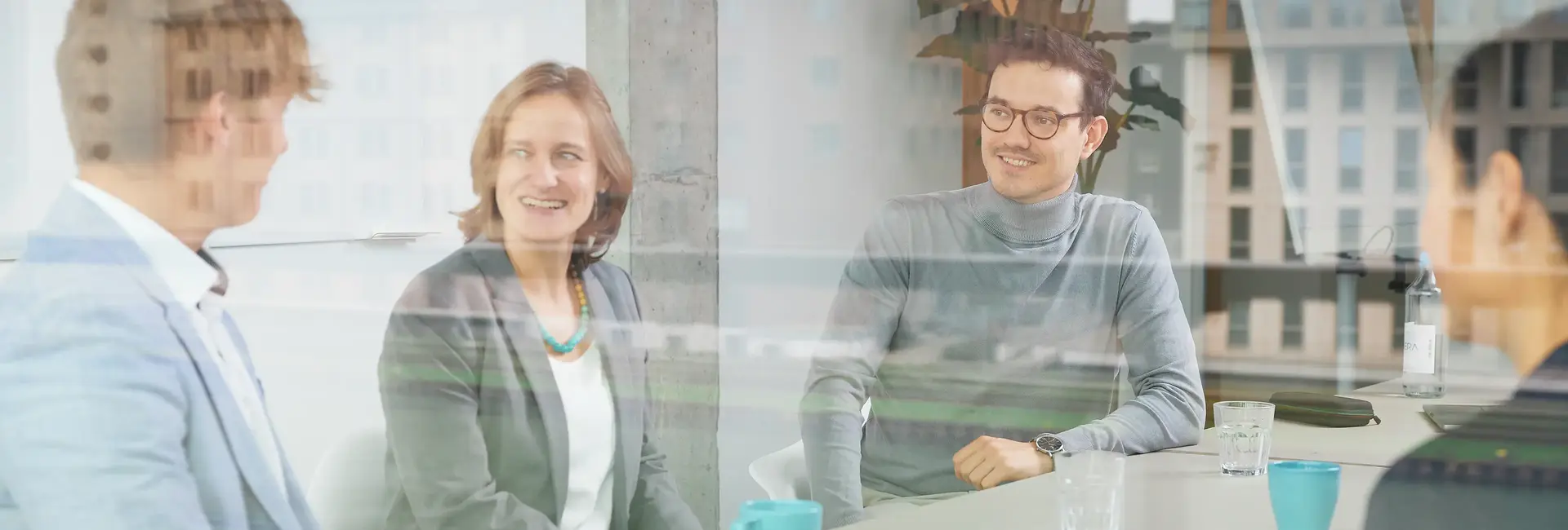 Mehrere Personen sitzen in einem modernen Büro und unterhalten sich lächelnd. Die Fensterspiegelung zeigt die Stadtlandschaft im Hintergrund.