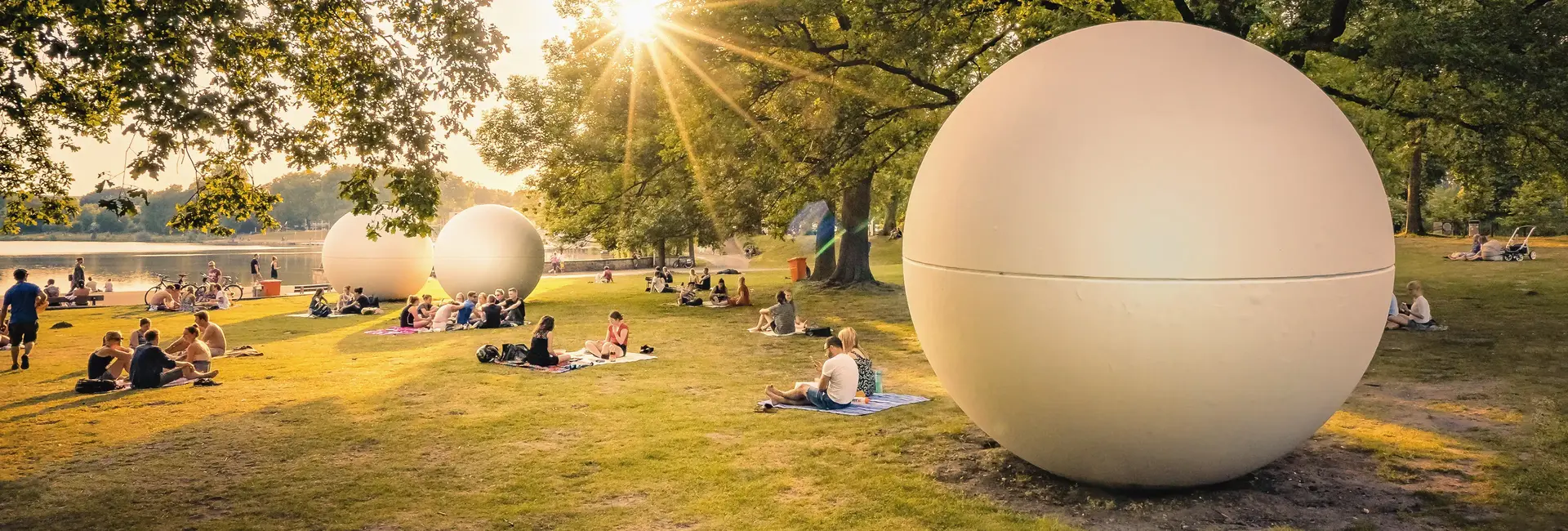 Menschen sitzen auf Picknickdecken im Grünen am Aasee in Münster, umgeben von großen kugelförmigen Kunstobjekten. Die Sonne strahlt durch die Bäume.