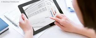 Eine Person nutzt ein Tablet zur Analyse von Geschäftsdaten mit Diagrammen und Tabellen, während auf dem Schreibtisch Unterlagen und ein Stift liegen.