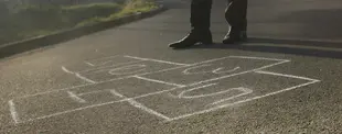 Das Bild zeigt ein Hüpfspiel mit Kreide auf Asphalt gezeichnet. Eine Person in Business-Schuhen steht daneben.