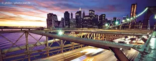 Das Bild zeigt eine beleuchtete Stadtansicht bei Sonnenuntergang, aufgenommen von einer Brücke mit vorbeifahrenden Autos, die Lichtspuren hinterlassen.
