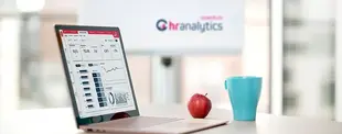 Ein Laptop mit einem HR-Analytics-Dashboard steht auf einem Schreibtisch, daneben eine blaue Tasse und ein Apfel. Im Hintergrund ist das noventum hr analytics Logo.