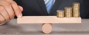 Ein Mann im Anzug balanciert eine Holz-Wippe mit Münzstapeln auf einem runden Holzstück, symbolisch für Finanz- oder Leistungsoptimierung.