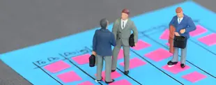 Miniaturfiguren in Business-Outfits stehen auf einem blauen Kanban-Board mit rosa Haftnotizen und diskutieren, während eine weitere Figur mit Aktenkoffer hinzukommt.
