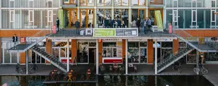 Ein modernes Gebäude mit einer Terrasse über dem Wasser, auf der Menschen bei einer Business-Veranstaltung stehen. Unten befinden sich Geschäfte und ein Café mit Sitzplätzen.