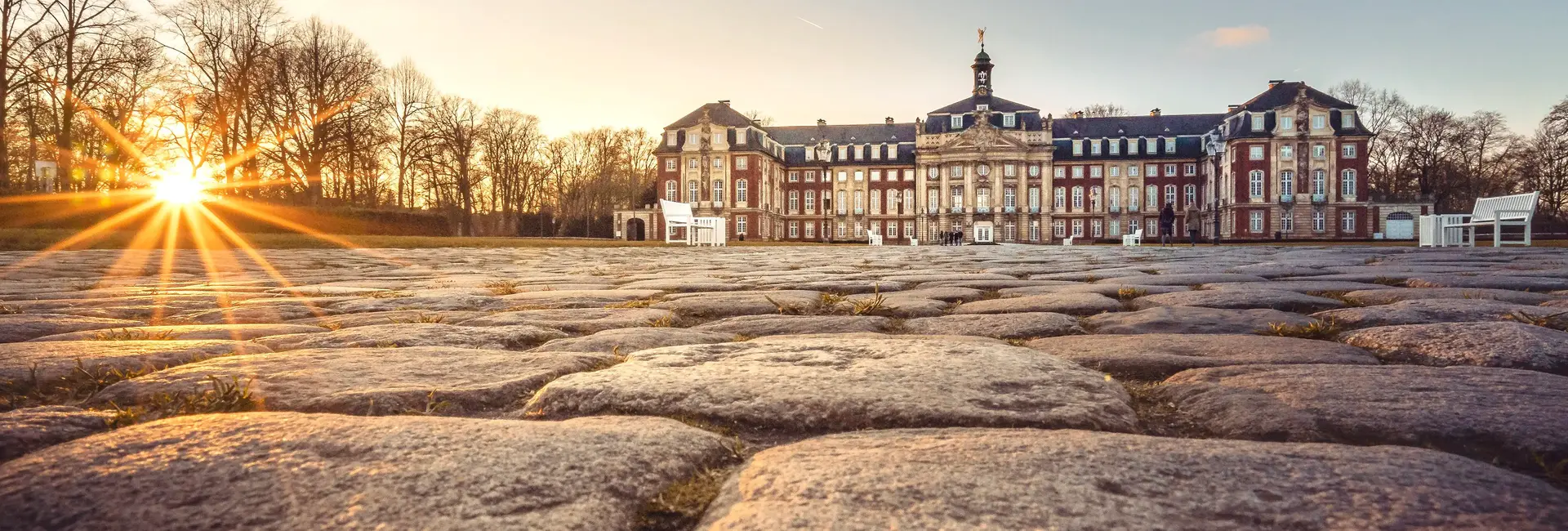 Das Bild zeigt das Schloss Münster mit gepflastertem Vorplatz bei tief stehender Sonne.