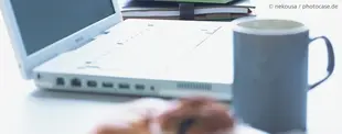 Das Bild zeigt einen Schreibtisch mit einem geöffneten Laptop, einer Kaffeetasse und einem Gebäckstück im Vordergrund, im Hintergrund sind Notizbücher gestapelt.