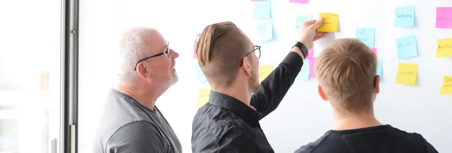 Drei Personen stehen vor einem Whiteboard mit bunten Notizzetteln. Eine Person zeigt auf eine Notiz, während die anderen aufmerksam zuhören und mitdiskutieren.