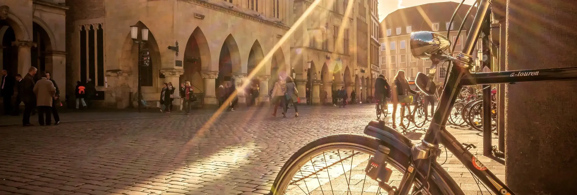Das Bild zeigt das historische Rathaus in Münster mit Passanten auf dem gepflasterten Prinzipalmarkt. Im Vordergrund lehnt ein Fahrrad an einer Säule.
