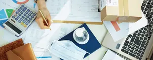Ein Schreibtisch mit Laptop, Dokumenten, Taschenrechner, Paket, Maske und Desinfektionsmittel und eine schreibende Hand.