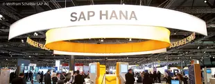 Ein großer SAP-HANA-Messestand mit leuchtendem Rundbanner auf einer Fachmesse, umgeben von zahlreichen Besuchern und digitalen Präsentationen.