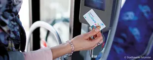 Eine Person hält eine Stadtwerke Münster PlusCard an ein Ticketlesegerät im Bus, um ihre Fahrt zu validieren. Der Bildschirm des Geräts zeigt eine Bestätigung an.
