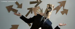 Eine Person im Anzug mit einer Papiertüte mit Fragezeichen über dem Kopf, umgeben von Pfeilen aus Pappe, symbolisiert Orientierungslosigkeit oder Entscheidungsdruck