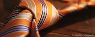 Das Bild zeigt eine orange gestreifte Krawatte mit einem Knoten auf einem Holztisch, symbolisch für Business, Formalität oder strategische Entscheidungen.