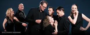 Das Bild zeigt sieben Personen in schwarzer Kleidung vor dunklem Hintergrund, die in verschiedenen theatralischen Posen interagieren, mit ausdrucksstarken Gesten und Mimik.