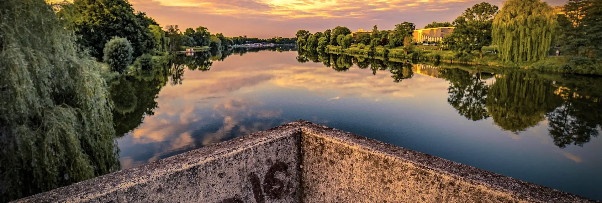 Der Aasee in Münster bei Sonnenuntergang, mit spiegelglatter Wasseroberfläche, die den Himmel reflektiert. Umgeben von grünen Bäumen und moderner Architektur.