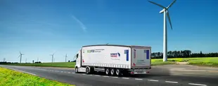 Ein Lkw mit Schmitz Cargobull Anhänger fährt auf einer Landstraße, umgeben von grünen Feldern und Windrädern.