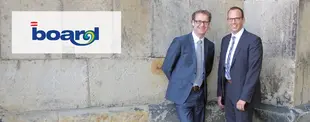 Zwei Männer in Anzügen stehen vor einer Steinwand und lächeln. Links im Bild ist das Logo von „Board“ zu sehen.