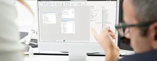 Ein Mann mit Brille zeigt auf einen Monitor, auf dem ein Datenmodell in Power BI zu sehen ist. Eine weitere Person steht daneben und betrachtet den Bildschirm