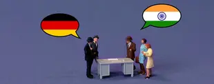 Miniaturfiguren stehen an einem Tisch und diskutieren. Sprechblasen mit den Flaggen Deutschlands und Indiens symbolisieren interkulturelle Kommunikation.