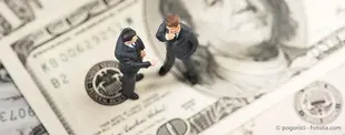 Das Bild zeigt zwei Miniaturfiguren in Business-Anzügen, die auf einem 100-Dollar-Schein stehen und diskutieren.