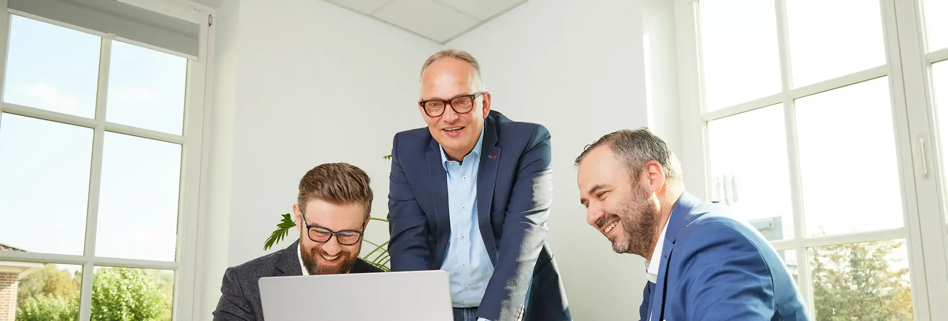 Drei Männer in Business-Kleidung lächeln und arbeiten gemeinsam an einem Laptop in einem lichtdurchfluteten Büro mit großen Fenstern und Blick ins Grüne.