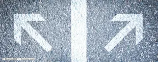 Zwei weiße Pfeile auf Asphalt zeigen in entgegengesetzte Richtungen, getrennt durch eine Mittellinie, symbolisch für Entscheidungen oder verschiedene Wege.