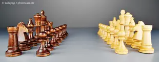 Das Bild zeigt zwei gegenüberstehende Schachfiguren-Sets in dunklem und hellem Holz, die sich auf einer ebenen Fläche gegenüberstehen, bereit für das Spiel.