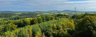 Panoramablick auf eine hügelige Landschaft mit dichten Wäldern, grünen Feldern, einem Sendemast und Windrädern unter einem teils bewölkten Himmel.