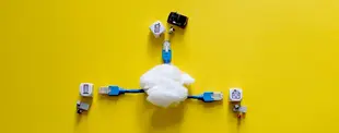Ein Wattebausch als „Cloud“ ist mit blauen Netzwerkkabeln verbunden, die zu Symbolwürfeln für Speicher, Dokumente und Daten führen, auf gelbem Hintergrund.