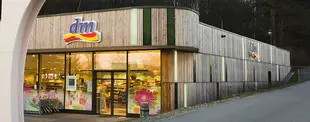 Ein dm-Markt mit moderner Holzfassade und großen Glasfenstern, durch die das beleuchtete Innere sichtbar ist. Einkaufswagen stehen vor dem Eingang