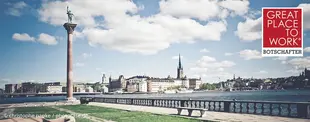Ein Blick auf eine historische Stadt am Wasser mit einer hohen Säule im Vordergrund. Oben rechts ist das Logo „Great Place to Work – Botschafter“ zu sehen.