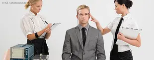 Ein Mann im Anzug sitzt mit neutralem Gesichtsausdruck, während zwei Frauen in Blusen und Krawatten ihn mit Klemmbrettern und Messgeräten untersuchen.