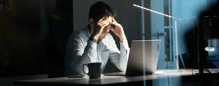 Ein Mann sitzt spät abends an einem Schreibtisch vor seinem Laptop, hält den Kopf in den Händen und wirkt erschöpft. Eine Lampe beleuchtet den dunklen Raum.