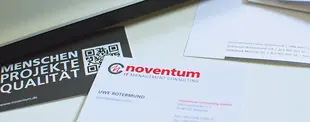 Visitenkarten und Geschäftspapiere von noventum consulting auf einem Schreibtisch. Ein schwarzes Kärtchen mit QR-Code trägt die Worte „MENSCHEN PROJEKTE QUALITÄT“.