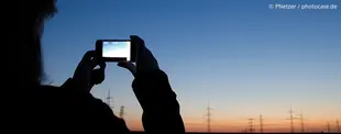 Eine Person fotografiert mit einem Smartphone einen Sonnenuntergang mit Strommasten im Hintergrund.