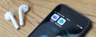 Ein Smartphone mit den Apps Microsoft Teams und SharePoint liegt auf einem Holztisch. Daneben befinden sich zwei kabellose weiße Ohrhörer.