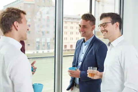 Vier Männer in Business-Kleidung stehen in einem modernen Büro vor großen Fenstern und unterhalten sich entspannt mit Getränken in der Hand, während sie lächeln.