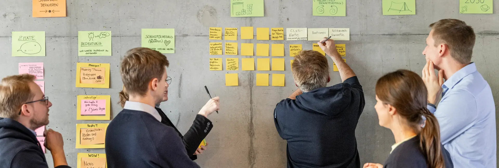 Fünf Personen stehen vor einer Wand mit bunten Klebezetteln, schreiben und diskutieren gemeinsam Ideen.