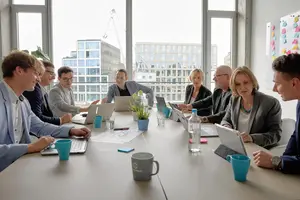 Eine Gruppe von Geschäftsleuten sitzt in einem modernen Büro an einem großen Tisch, arbeitet mit Laptops und diskutiert konzentriert. Helle Fensterfront im Hintergrund.