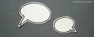 Das Bild zeigt zwei ausgeschnittene Sprechblasen auf grauem Stoffhintergrund, symbolisch für Kommunikation, Dialog oder kreative Ideenentwicklung.