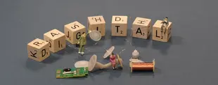 Scrabble-Steine formen das Wort „DIGITAL“. Miniaturfiguren arbeiten mit Zahnrädern, Leiterplatte und Draht, symbolisieren Digitalisierung und Technik.