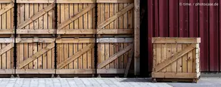 Mehrere gestapelte Holzkisten vor einer roten Metallwand, eine einzelne Kiste steht separat daneben auf einem gepflasterten Boden.