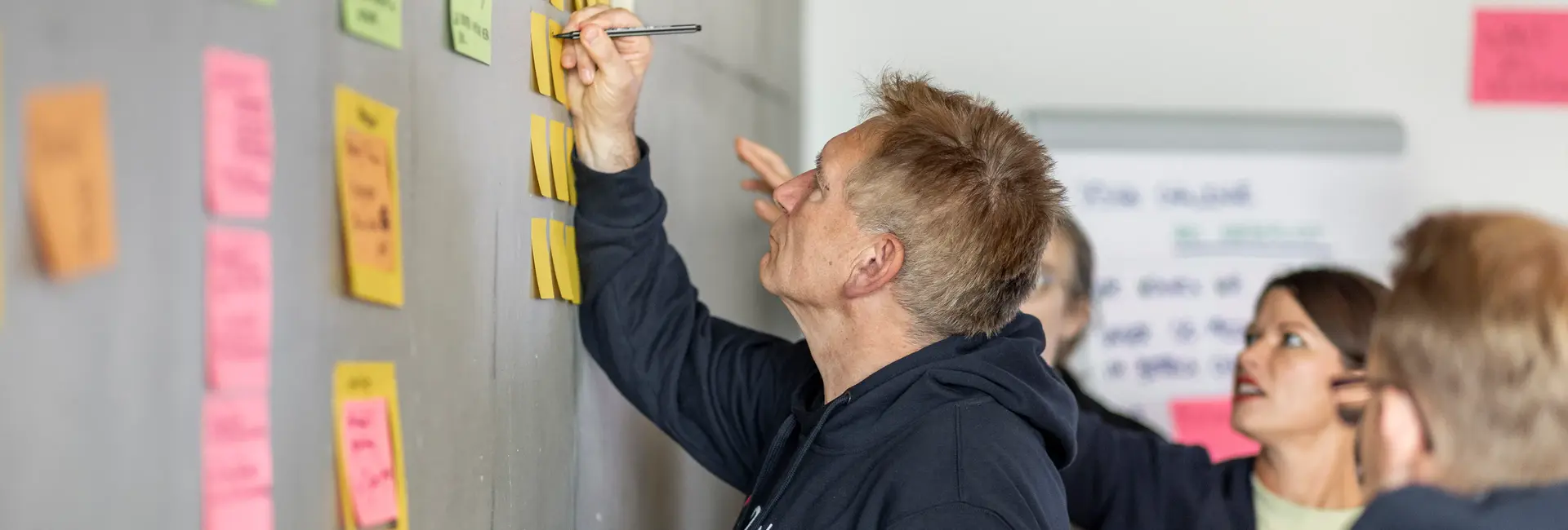 Uwe Rotermund schreibt auf eine Haftnotiz an einer Wand voller Klebezettel. Mehrere Personen im Hintergrund diskutieren und beteiligen sich an der Ideensammlung.