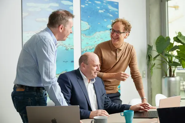 Drei Männer in Business-Kleidung lachen und diskutieren an einem Tisch mit Laptops und Kaffeetassen in einem modernen Büro mit großen Pflanzen und Wandbildern.