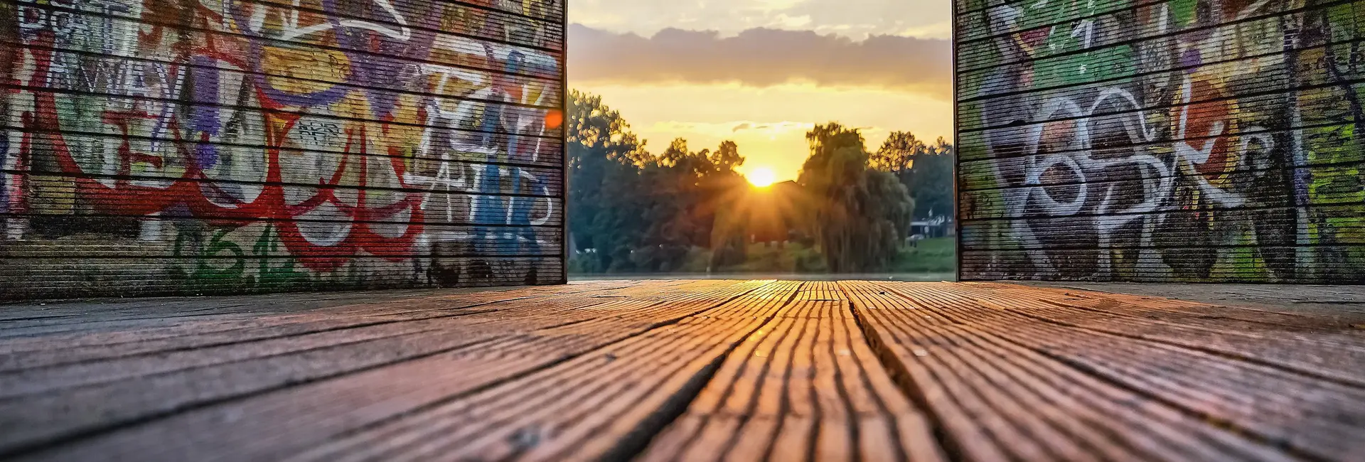 Holzplattform am Münsteraner Aasee mit Graffiti-besprühten Wänden öffnet sich zu einer Landschaft mit Bäumen und Sonnenuntergang im Hintergrund.