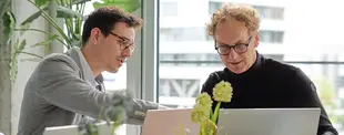 Zwei Männer arbeiten konzentriert an Laptops in einem modernen Büro mit Pflanzen und großen Fenstern.